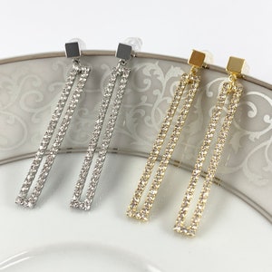 May include: Three pairs of dangle earrings with a rectangular shape. The earrings are made of silver, gold, and rose gold metal and are adorned with clear rhinestones.