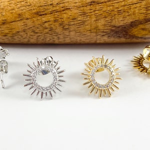 May include: A pair of earrings with a sunburst design. The earrings are available in silver and gold. Each earring features a circle of small clear stones surrounded by a ring of pointed rays.