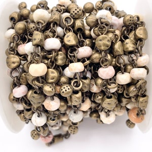 May include: A close-up of a chain of beads and metal charms. The beads are white, pink, and beige, and the charms are antique brass. The chain is coiled up on a white surface.