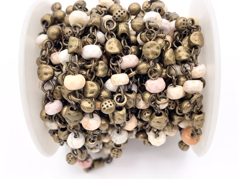 May include: A close-up of a chain of beads and metal charms. The beads are white, pink, and beige, and the charms are antique brass. The chain is coiled up on a white surface.