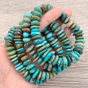 May include: Strands of turquoise gemstone beads in shades of blue, green, and brown. The oval-shaped beads are strung together, creating a textured accessory. The gemstones are held in a hand. The beads are measured in centimetres.