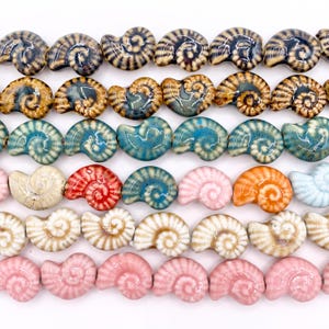 May include: A collection of ceramic beads shaped like nautilus shells, arranged in horizontal rows. The beads feature a glossy finish and a variety of colours, including shades of blue, brown, pink, orange, red, and cream, with some displaying striped patterns.