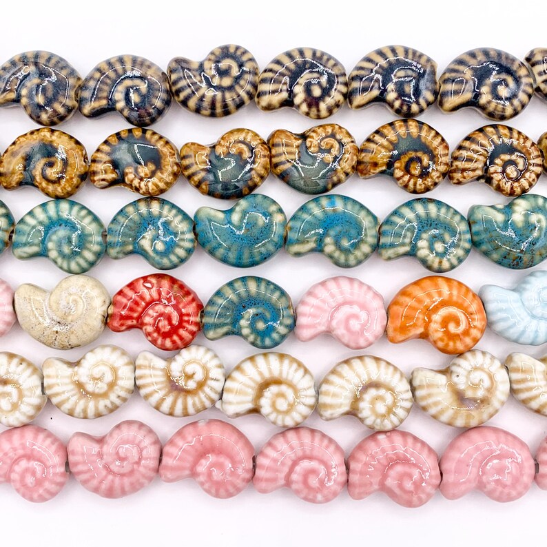 May include: A collection of ceramic beads shaped like nautilus shells, arranged in horizontal rows. The beads feature a glossy finish and a variety of colours, including shades of blue, brown, pink, orange, red, and cream, with some displaying striped patterns.