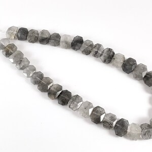 May include: A strand of faceted, smoky quartz beads. The beads are a mix of light and dark gray, with some showing a hint of clear quartz. The beads are strung on a clear elastic cord.