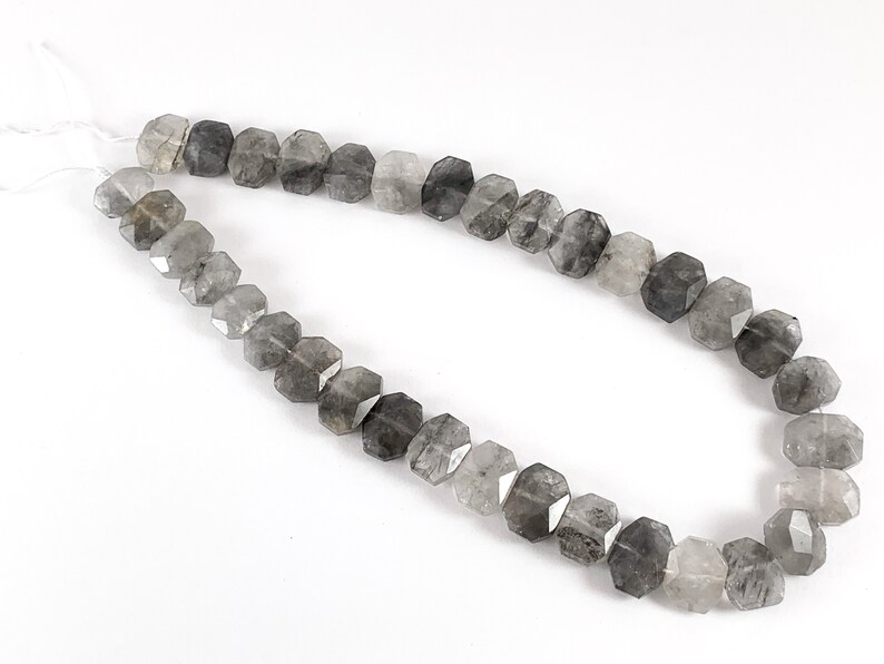 May include: A strand of faceted, smoky quartz beads. The beads are a mix of light and dark gray, with some showing a hint of clear quartz. The beads are strung on a clear elastic cord.