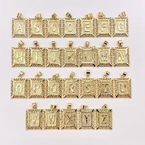 May include: A collection of gold-colored initial pendants. Each pendant features a square frame with a decorative filigree design. The letters A through Z are displayed in a bold, capital font within the frames. The pendants are designed for necklaces.