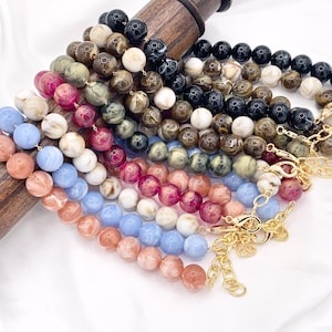 May include: A collection of beaded necklaces in various colors, including black, white, pink, blue, and brown. Each necklace features large, round beads and gold-colored chain and clasp closures. The necklaces are displayed on a wooden dowel against a white background.