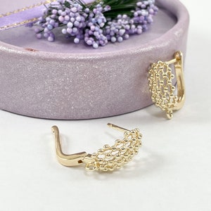 May include: A pair of gold-toned hoop earrings with a delicate, textured design. The earrings are made of small, round beads that are woven together to create a mesh-like pattern.