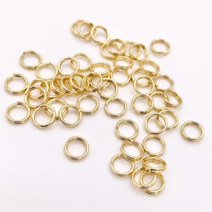 May include: A collection of numerous small, circular gold-toned metal jump rings scattered across the frame. Each individual ring features a split design, allowing for opening and closing, with a polished, reflective surface finish throughout.
