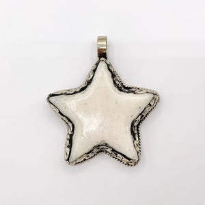 May include: A star-shaped pendant with a white center and a silver-colored, ornate border. The pendant has a loop at the top for a chain. The design is symmetrical and the star has five points.