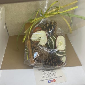 May include: A clear plastic bag filled with dried apple slices, cinnamon sticks, star anise, and pine cones. The bag is tied with a green and brown ribbon. A small card with the text "Kindness by the lovely people at HOMESPUN NATURAL HOME FRAGRANCES Thank you for your continued support" is visible in the bottom right corner of the image.