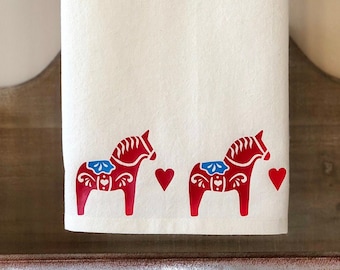 May include: Two red Swedish Dala horses with blue and white details are printed on a white kitchen towel. The towel is hanging on a rustic wooden and metal shelf.