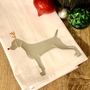 May include: A white kitchen towel with a gray Weimaraner dog silhouette printed on it. The dog has red antlers and a red nose, making it look like a reindeer. The towel is folded and lying on a wooden surface with Christmas ornaments in the background.