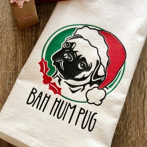 May include: White kitchen towel with a black and white illustration of a pug wearing a Santa hat. The pug is in a green circle with red holly and the text "Bah Hum Pug".