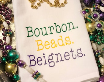 May include: White kitchen towel with the text "Bourbon. Beads. Beignets." in green, yellow, and purple lettering. The towel is surrounded by purple, gold, and green Mardi Gras beads.