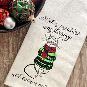 May include: A white kitchen towel with a black and green illustration of a mouse wearing a green and black sweater and a red scarf. The text "Not a creature was stirring not even a mouse." is printed on the towel.