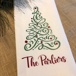 K&ouml;nnte beinhalten: Ein wei&szlig;es K&uuml;chentuch mit einem gr&uuml;nen und roten Weihnachtsbaumdesign und dem Text "The Parliers" in Rot.