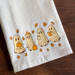 以下が含まれることがあります： 白いハロウィーンをテーマにしたキッチンタオル。4体の漫画の幽霊が描かれており、カボチャ、コーヒー、本を持っています。幽霊はベージュ色で茶色の輪郭があり、オレンジ色の葉が周りにあります。装飾的で、お祝いのアイテムです。