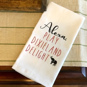 May include: White kitchen towel with the text "Alexa, Play Dixieland Delight" in red lettering and a black silhouette of an elephant.