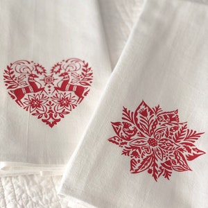 May include: Two white kitchen towels with red holiday designs. One towel features a heart-shaped design with two reindeer and floral accents. The other towel features a red poinsettia design with floral accents.