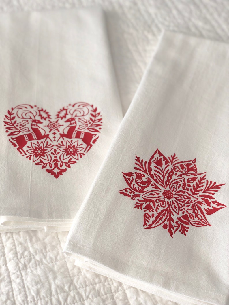 May include: Two white kitchen towels with red holiday designs. One towel features a heart-shaped design with two reindeer and floral accents. The other towel features a red poinsettia design with floral accents.