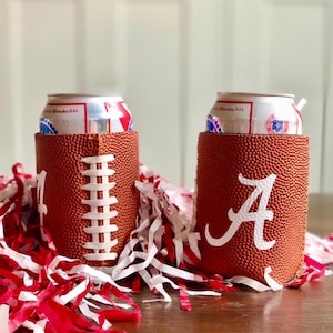 May include: Two brown football-shaped can coolers with white stitching and white lettering. One cooler has the letter "A", and the other has the white stitching of a football. Both coolers hold a can of beer. Red and white streamers are in the foreground.