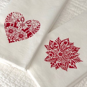 May include: Two white kitchen towels with red holiday designs. One towel features a heart-shaped design with two reindeer and floral elements. The other towel features a red poinsettia with floral elements.