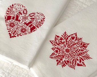 May include: Two white kitchen towels with red holiday designs. One towel features a heart-shaped design with two reindeer and floral elements. The other towel features a red poinsettia with floral elements.