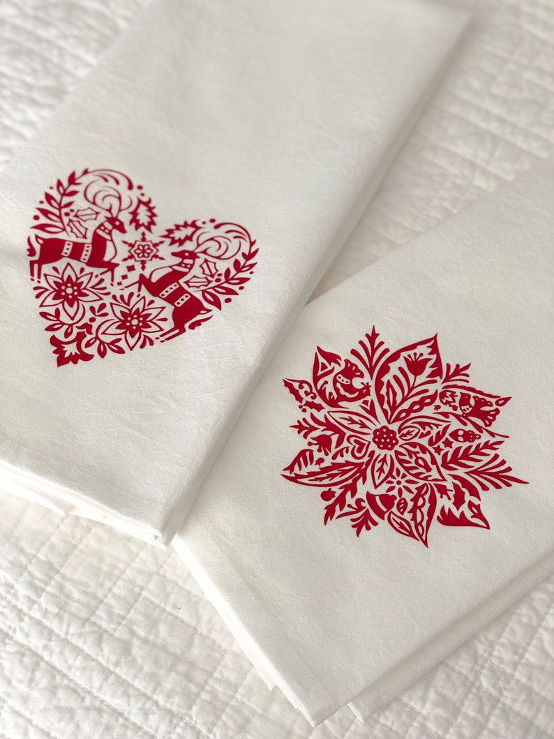 May include: Two white kitchen towels with red holiday designs. One towel features a heart-shaped design with two reindeer and floral elements. The other towel features a red poinsettia with floral elements.
