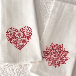May include: Two white kitchen towels with red embroidered designs. One towel features a heart-shaped design with two reindeer inside. The other towel features a red poinsettia design.