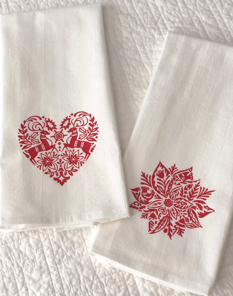 May include: Two white kitchen towels with red embroidered designs. One towel features a heart-shaped design with two reindeer inside. The other towel features a red poinsettia design.