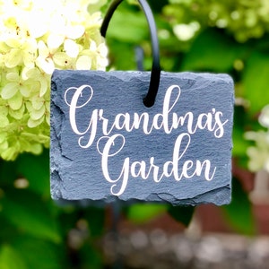 May include: A black slate garden marker with white text that reads "Grandma's Garden". The marker is attached to a black metal stake.
