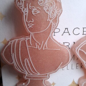 May include: Pink resin earrings featuring a bust of a classical sculpture with white outline details. The earrings have a gold-colored hoop at the top for hanging. The design is a classical art theme.