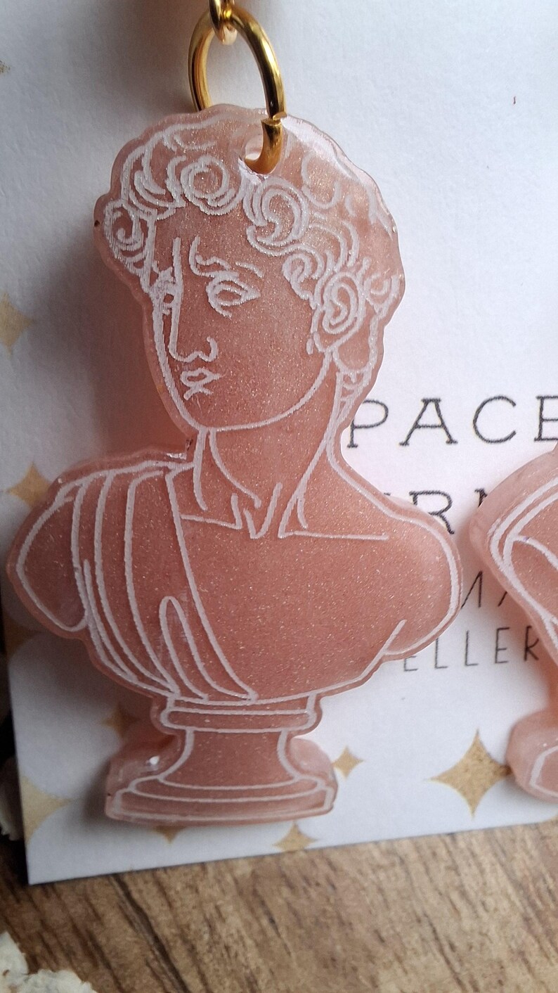 May include: Pink resin earrings featuring a bust of a classical sculpture with white outline details. The earrings have a gold-colored hoop at the top for hanging. The design is a classical art theme.