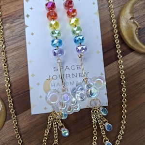 May include: A pair of handmade earrings with a rainbow gradient of faceted beads, gold chains, and iridescent bubble-shaped charms. The earrings are attached to a card that reads "SPACE JOURNEY HANDMADE JEWELRY".