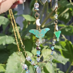 May include: A decorative glasses chain featuring a gold-toned metal link chain adorned with translucent mint green moth charms, small white crescent moon pendants, and a variety of clear, iridescent, and light blue spherical glass beads.