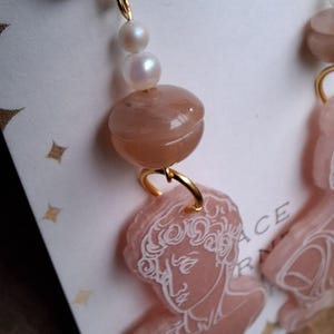 May include: Close-up of a pair of gold-toned earrings. Each earring features a lever back closure, a string of pearls, a peach-colored bead, and a pink pendant with a white engraved classical bust design. The earrings are on a white background.