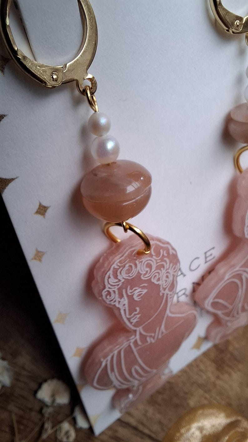 May include: Close-up of a pair of gold-toned earrings. Each earring features a lever back closure, a string of pearls, a peach-colored bead, and a pink pendant with a white engraved classical bust design. The earrings are on a white background.