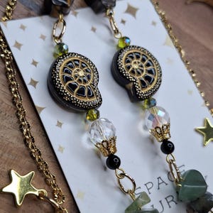 May include: A pair of dangle earrings featuring brown tiger eye beads, black and gold ammonite-shaped charms, faceted clear crystal beads, small green beads, and dangling green stone chips. The earrings are attached to a white display card.