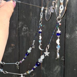 May include: A pair of round-framed glasses featuring a decorative beaded chain. The chain is composed of dark blue, iridescent, and clear beads, interspersed with silver-toned metal star and crescent moon charms, attached to the glasses with loops.