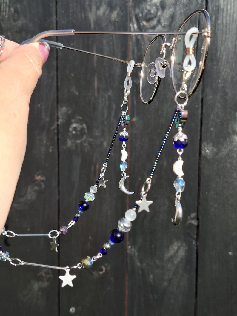 May include: A pair of round-framed glasses featuring a decorative beaded chain. The chain is composed of dark blue, iridescent, and clear beads, interspersed with silver-toned metal star and crescent moon charms, attached to the glasses with loops.