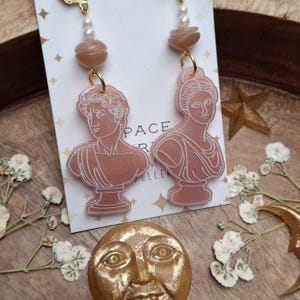May include: A pair of pink earrings featuring classical bust designs. Each earring has a gold-colored hoop, a small cluster of beads, and a bust of a person. The earrings are displayed on a white card with the text "PACE R M VELLER".