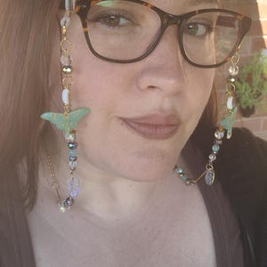 May include: Eyeglass chain with a gold-coloured chain, clear and iridescent beads, and a light green moth pendant. The chain is attached to tortoiseshell glasses. The glasses are worn by a person.