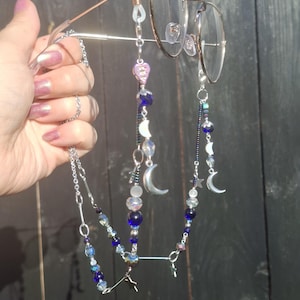 May include: A silver-toned eyeglass chain featuring a variety of beads in shades of blue, clear, and iridescent white. The chain includes metal charms shaped like crescent moons and stars, attached to the frames with clear silicone loops.