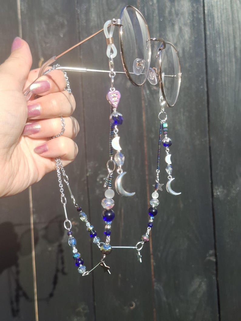 May include: A silver-toned eyeglass chain featuring a variety of beads in shades of blue, clear, and iridescent white. The chain includes metal charms shaped like crescent moons and stars, attached to the frames with clear silicone loops.