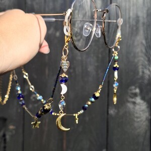May include: A gold-toned glasses chain featuring a variety of charms including a crescent moon, a heart, and small stars. The chain is decorated with dark blue, clear, and metallic beads, and it attaches to the frames with clear silicone loops.