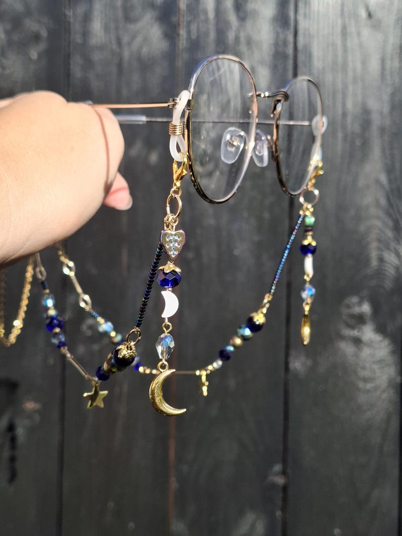 May include: A gold-toned glasses chain featuring a variety of charms including a crescent moon, a heart, and small stars. The chain is decorated with dark blue, clear, and metallic beads, and it attaches to the frames with clear silicone loops.