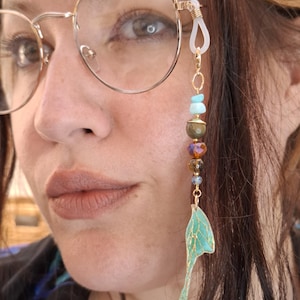 May include: A decorative glasses chain attachment featuring a series of beads in shades of turquoise, brown, and amber, finished with a gold-toned metal wing charm. The accessory connects to the temple of gold-rimmed eyeglasses via a clear silicone loop.
