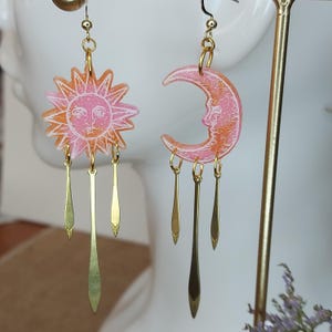 May include: A pair of dangle earrings featuring a pink and orange sun and moon design with gold accents. The earrings have three dangling gold charms on each side.