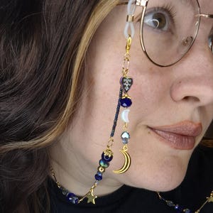 May include: A gold-toned eyeglass chain featuring a variety of blue beads, iridescent heart-shaped charms, a crescent moon charm, and a small star charm. The chain attaches to the frames with white silicone loops and gold-toned metal connectors.